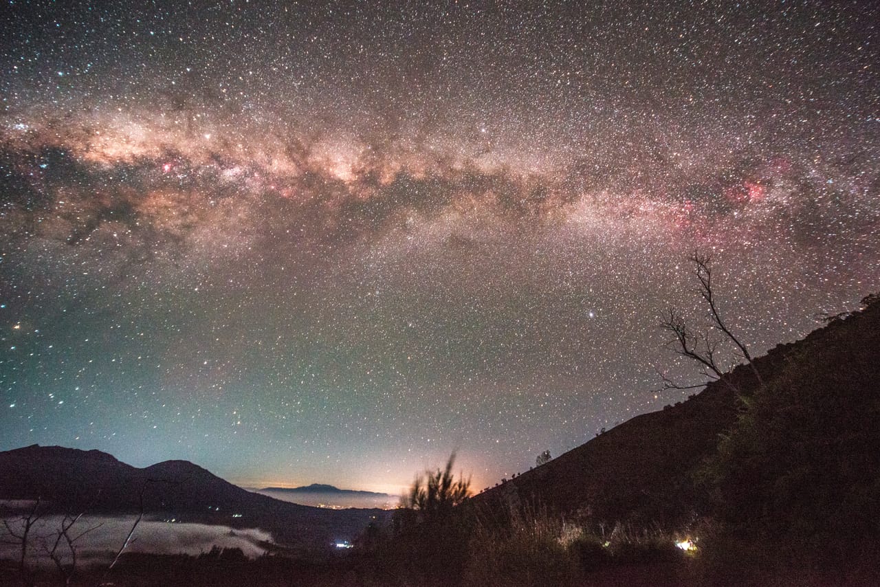 银河像一道拱横过夜空，下方是云海与火山的轮廓，山坡上立着一棵枯树。